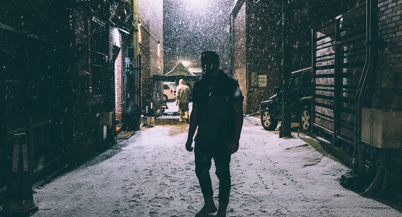 Lone figure walking out of snowy alley, flakes lit