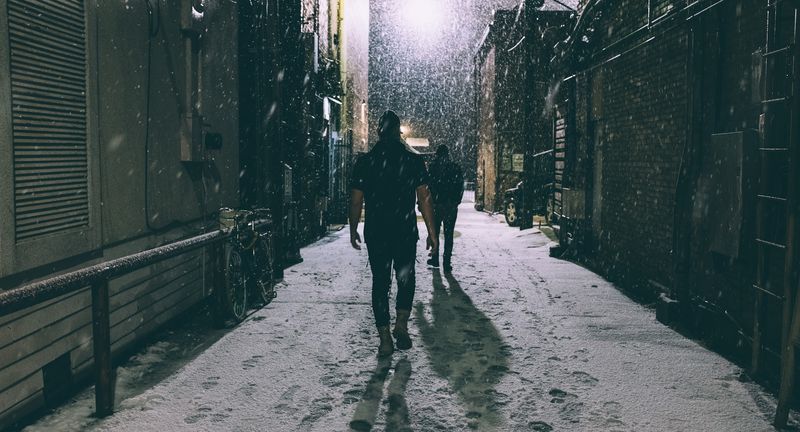 Two figures walk into falling snow down a backlit alley