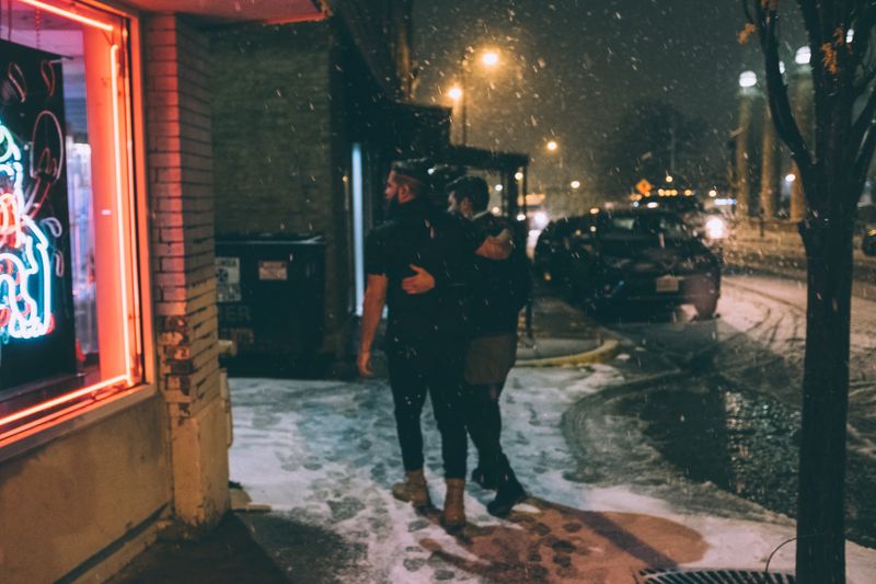 Pair walking snowy street past neon window at night