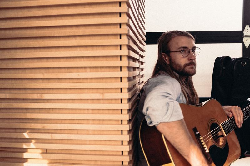 Glasses-wearing guitarist against warm wood slats, side light