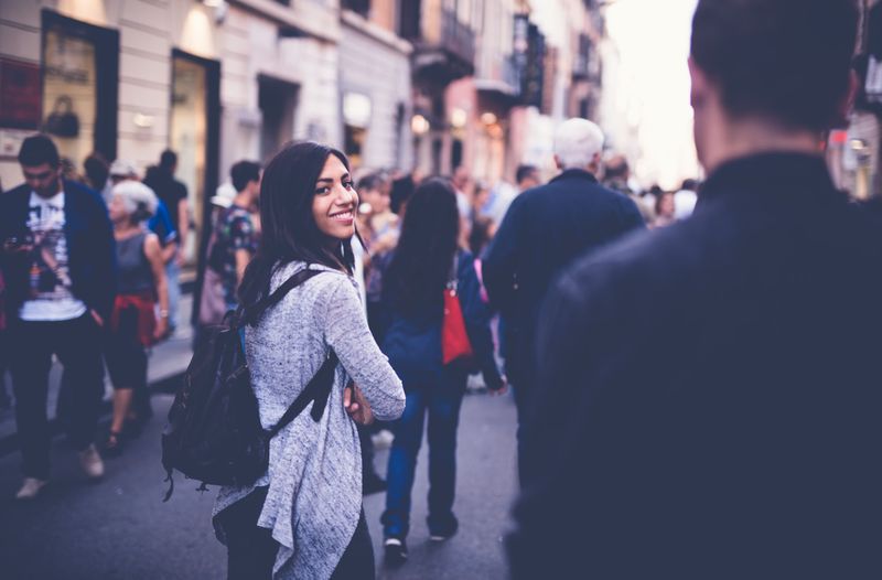 Glances back smiling through a crowded street, shoulder blurred in front