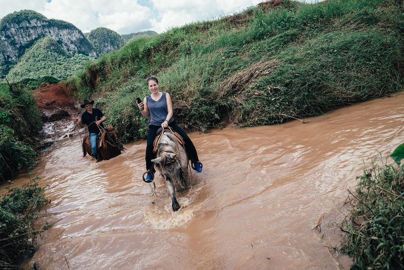 Riders crossing a muddy river, phone held up, green karst hills