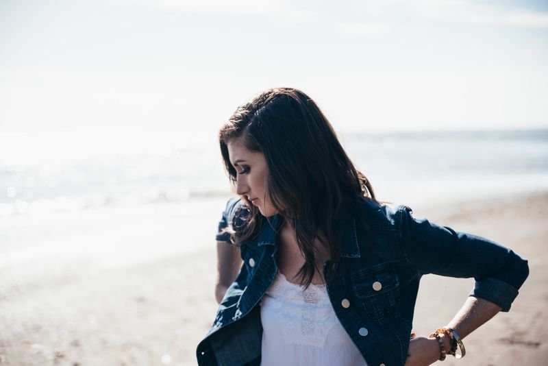 Denim jacket, hand at her hip, eyes down, bright sea behind