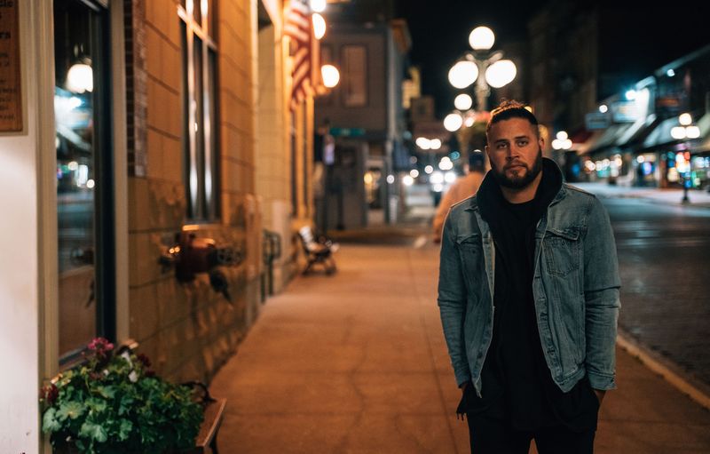 Man in a denim jacket on a night sidewalk, warm streetlights and flowers