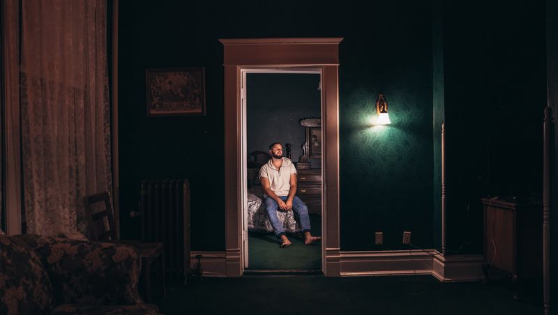 A man sits on a bed in a far doorway, a dark green room, one wall lamp