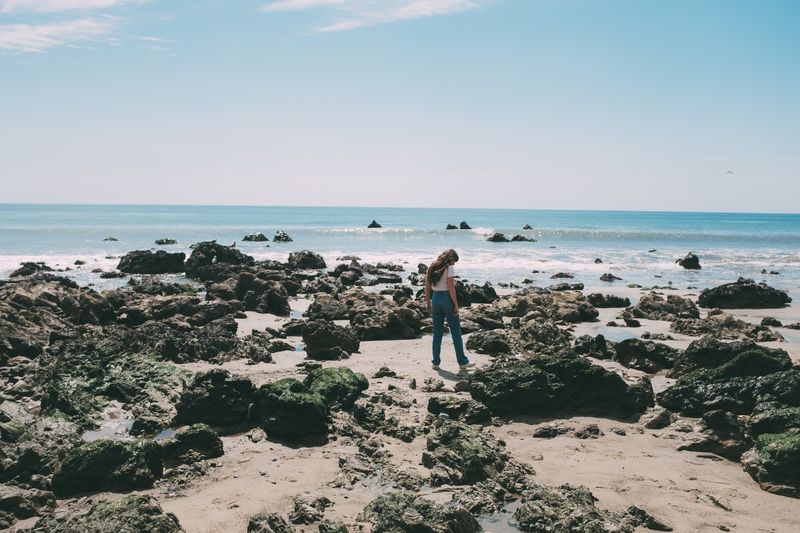 A small figure on a tide-pool beach, head down, rocks and surf behind
