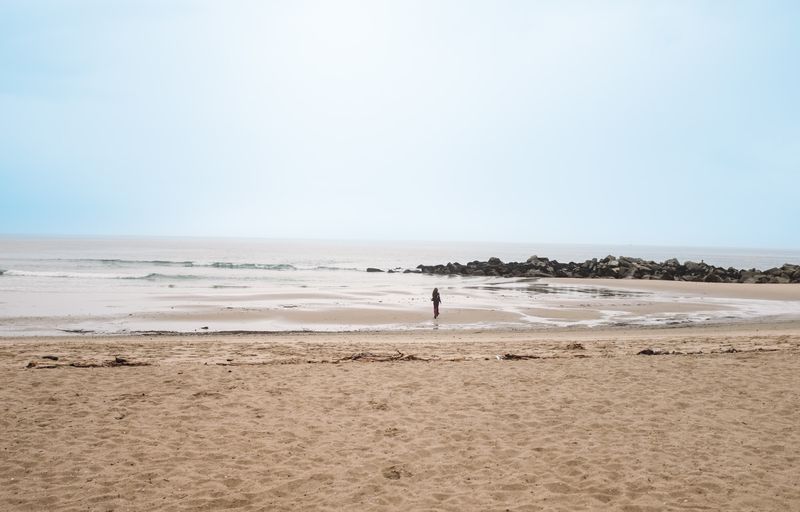A wide empty beach, one tiny figure at the waterline, flat pale sky