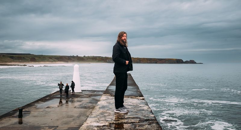 Tall figure on the pier, white marker, tiny walkers behind