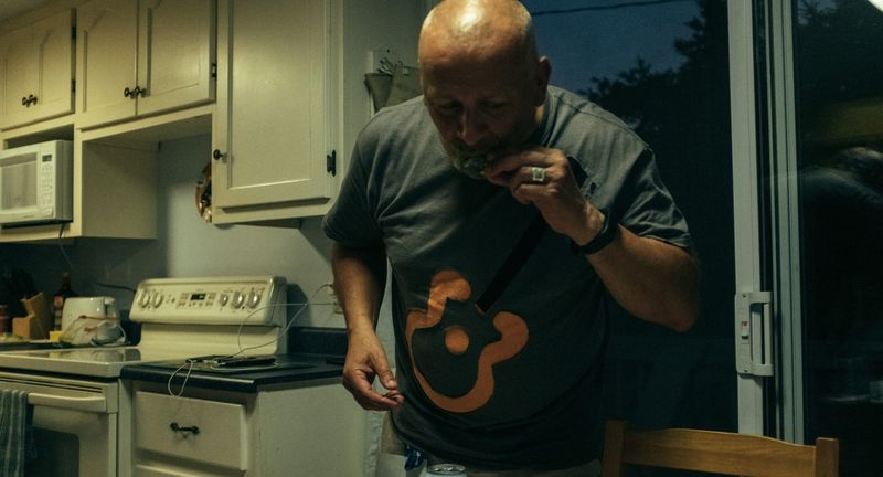 A bald man plays a harmonica in a kitchen, a guitar on his tee