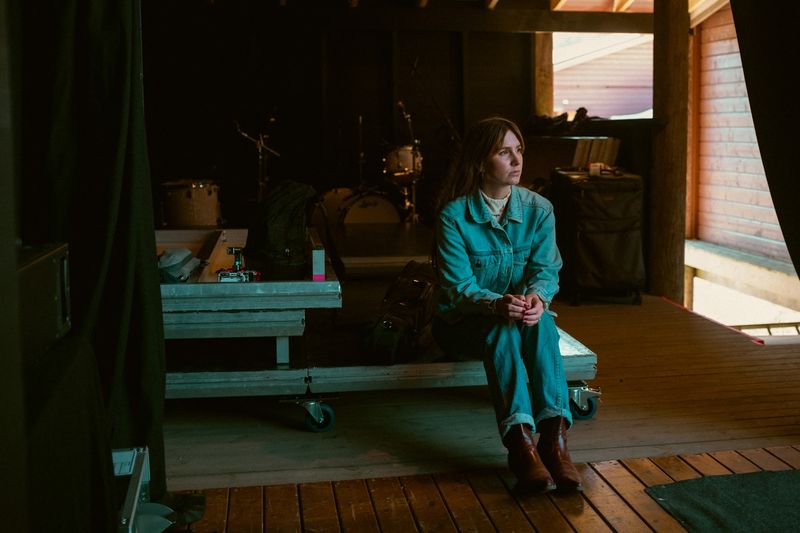 Woman in a denim jumpsuit sits on a flight case backstage, looking off
