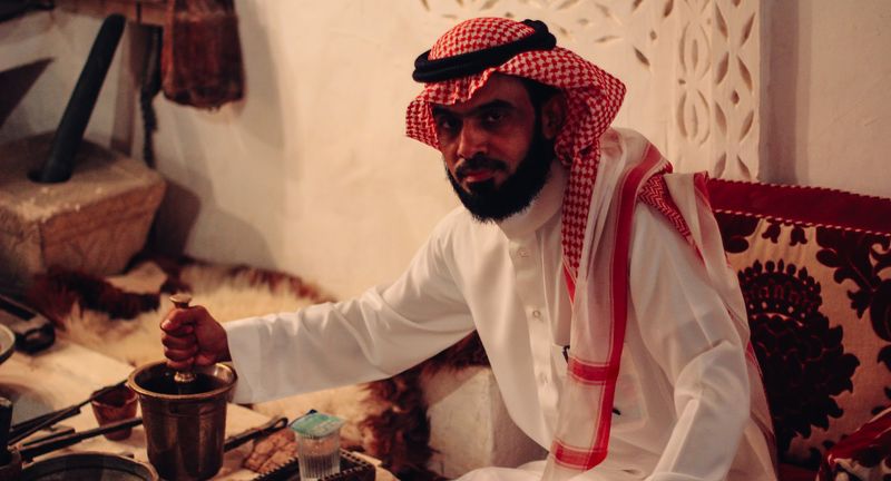 Red-checked headscarf, pestle in hand, direct gaze, warm room
