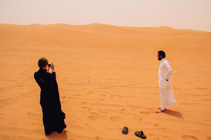 Black abaya and white thobe on orange dunes, sandals left behind