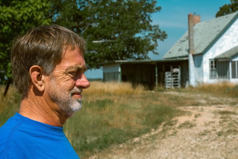 A man in a bright blue tee in profile, a pale farmhouse soft behind him
